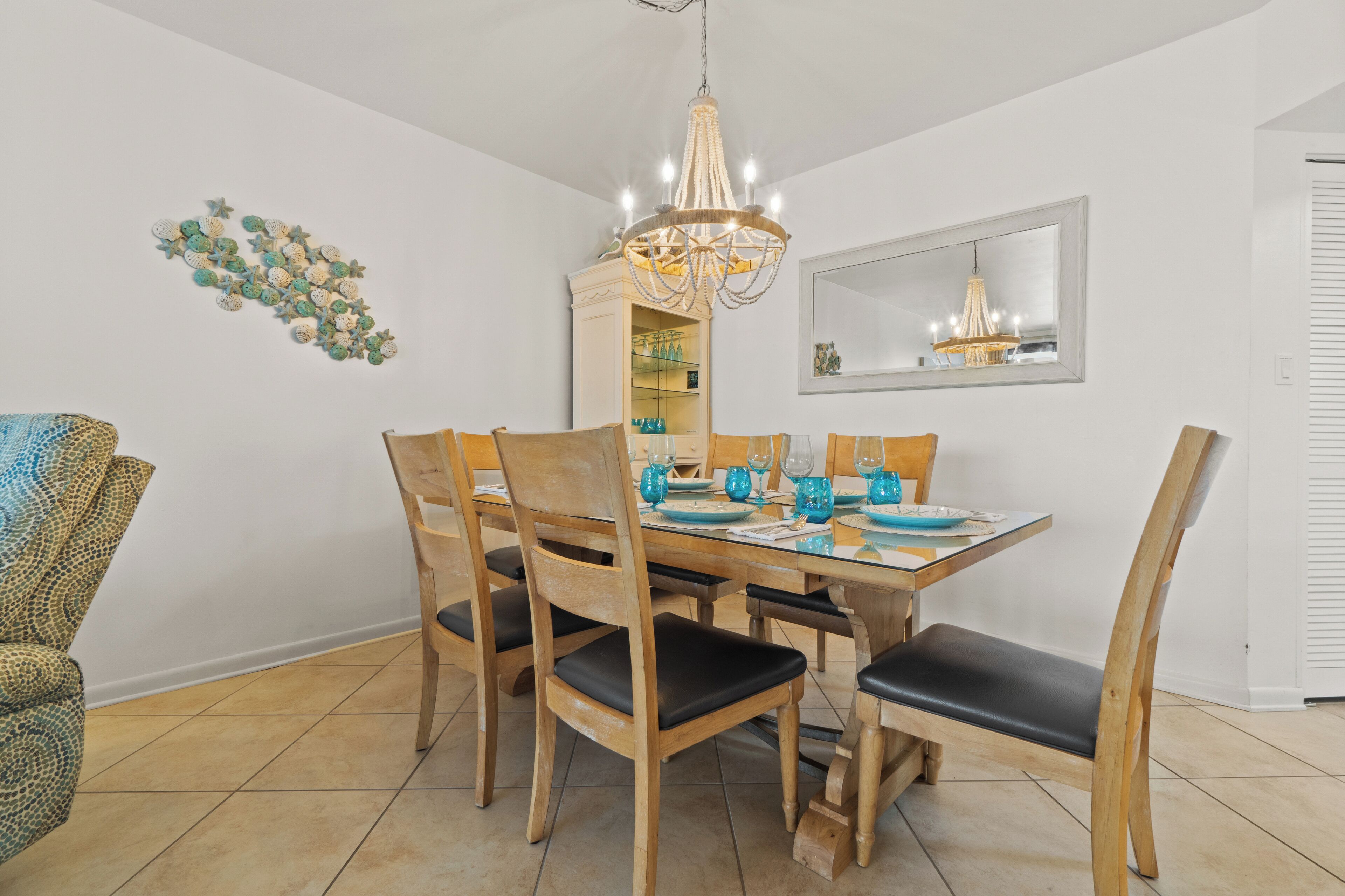 A dining area with a wooden table set for six, surrounded by matching chairs. A chandelier hangs above, and a decorative wall piece is visible to the left.