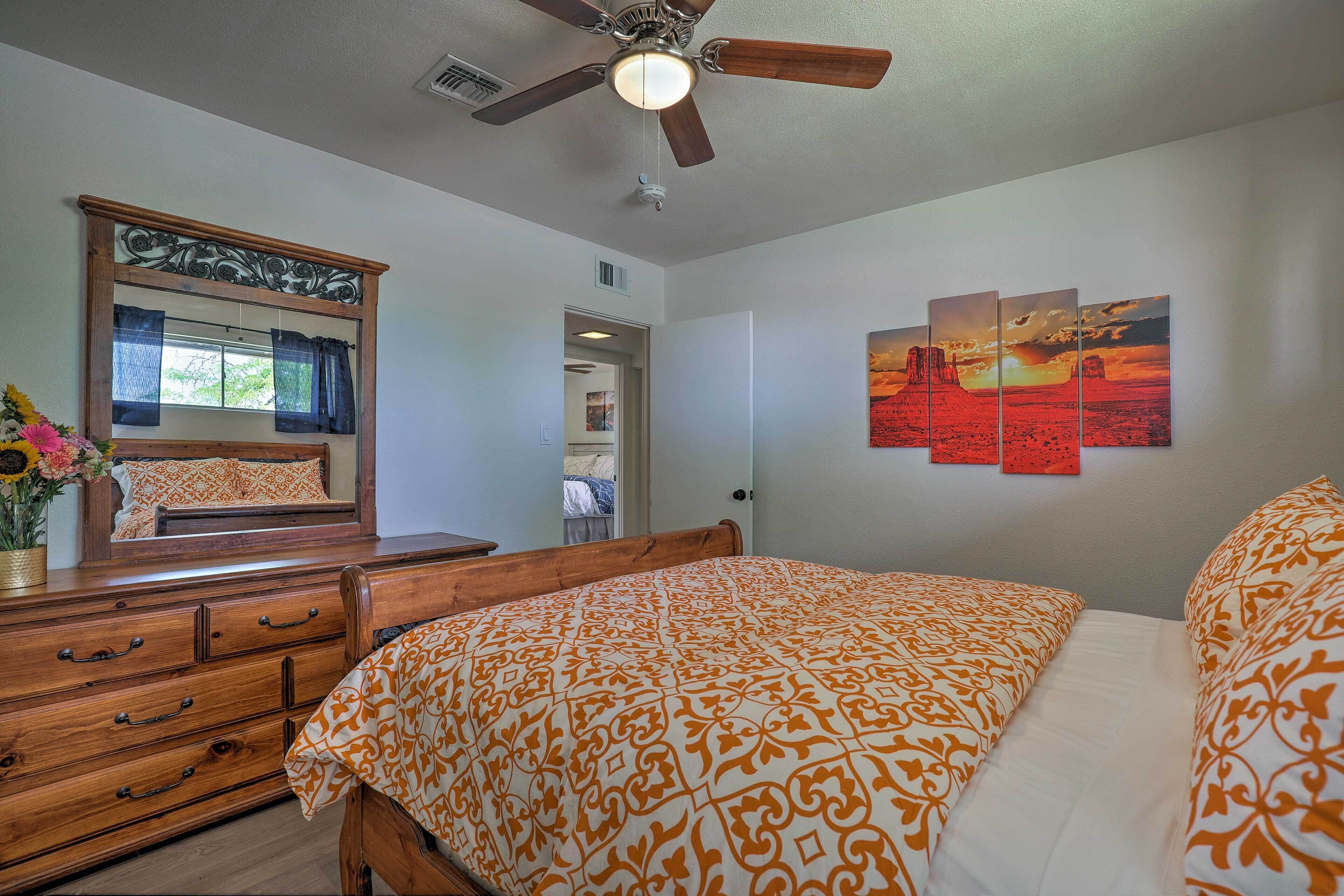 Un dormitorio con cama, cómoda y ventilador de techo. Se ve una ventana con cortinas y un espejo sobre la cómoda.
