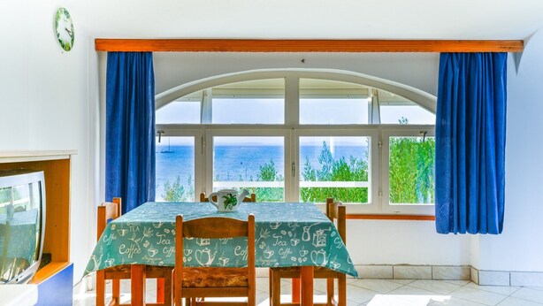 A dining area with a table set for four, a clock on the wall, and a view of the sea through large windows. - Summer Dream - One Bedroom Apartment with Sea View (Privlaka)