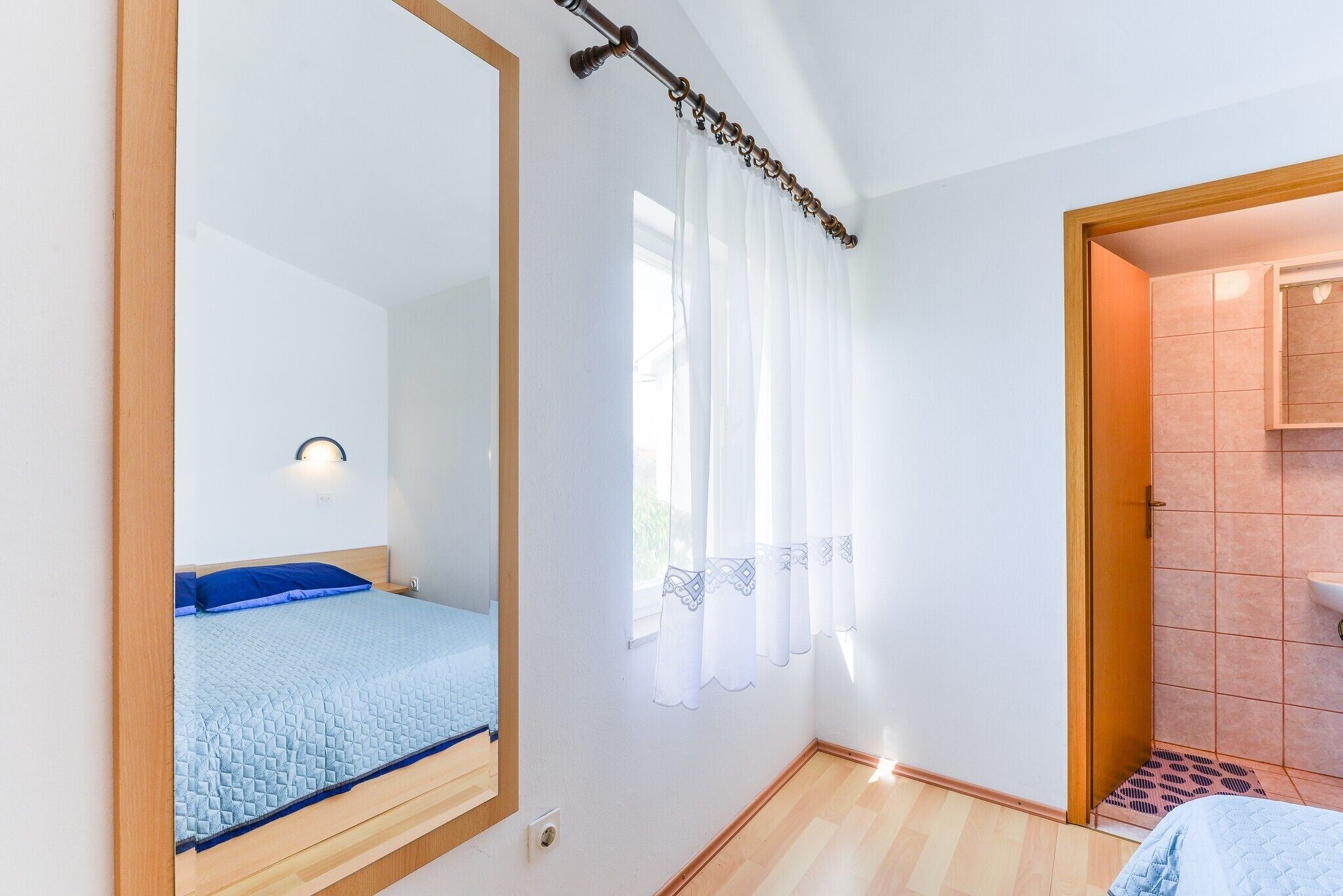 Quarto com cama, espelho, janela com cortinas e banheiro visível por uma porta aberta.