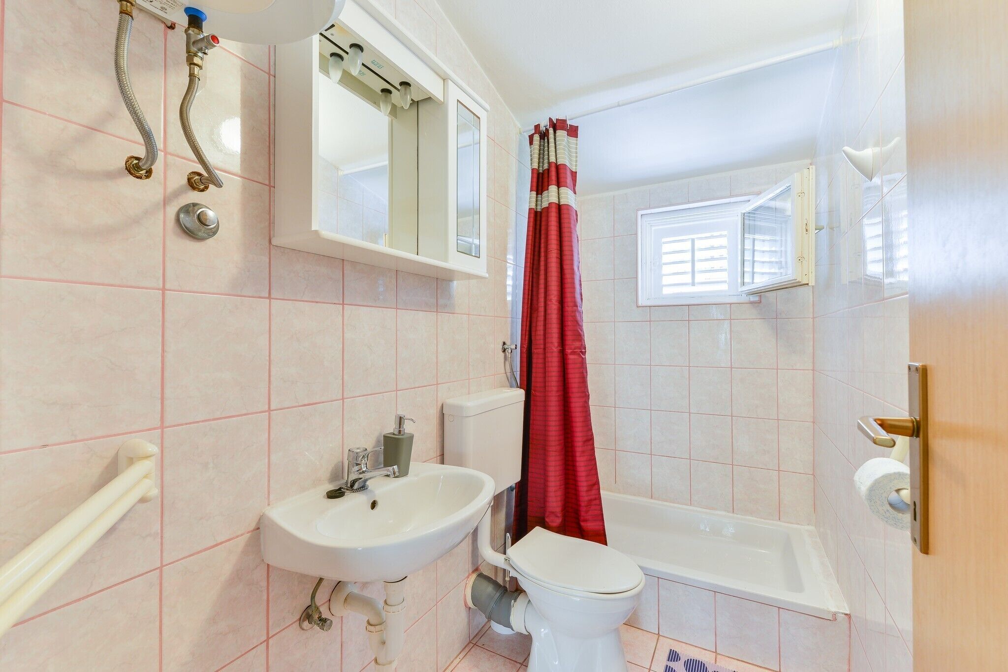 Une salle de bain avec un lavabo, des toilettes et une baignoire, tous de couleur blanche. Il y a une fenêtre avec un rideau rouge, et un miroir au-dessus du lavabo.