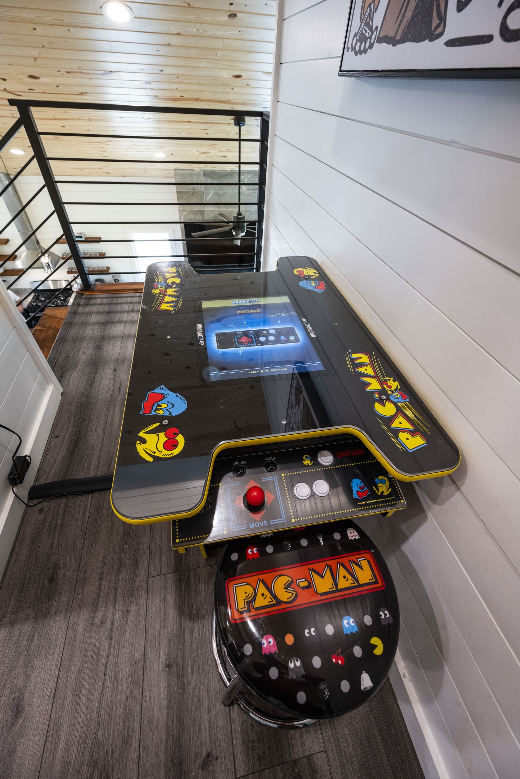 A retro arcade machine with a joystick and button panel, located in a hallway with wooden flooring and a metal railing.