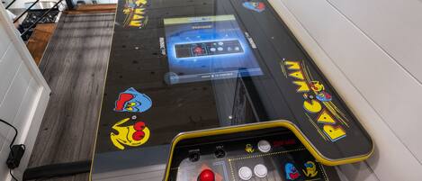 A retro arcade machine with a joystick and button panel, located in a hallway with wooden flooring and a metal railing.