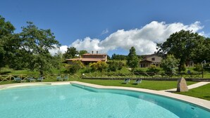 A swimming pool with lounge chairs, surrounded by a grassy area and stone walls, with houses in the background. - Villa Nocciola "Lovely cottage in the nature" (Roccalbegna)
