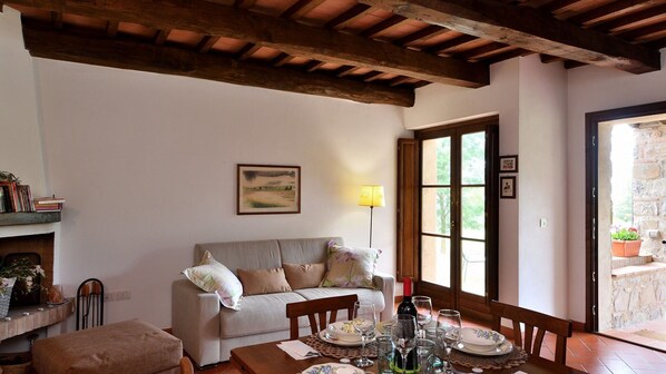 Un salon chaleureux avec un canapé, une table à manger pour deux personnes et une grande fenêtre donnant sur un paysage verdoyant.