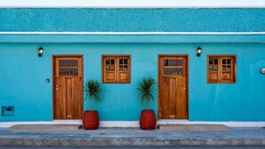 Un bâtiment doté de trois portes et fenêtres en bois, chacune étant munie d’une grille métallique. Deux grands pots de plantes sont placés devant les portes.