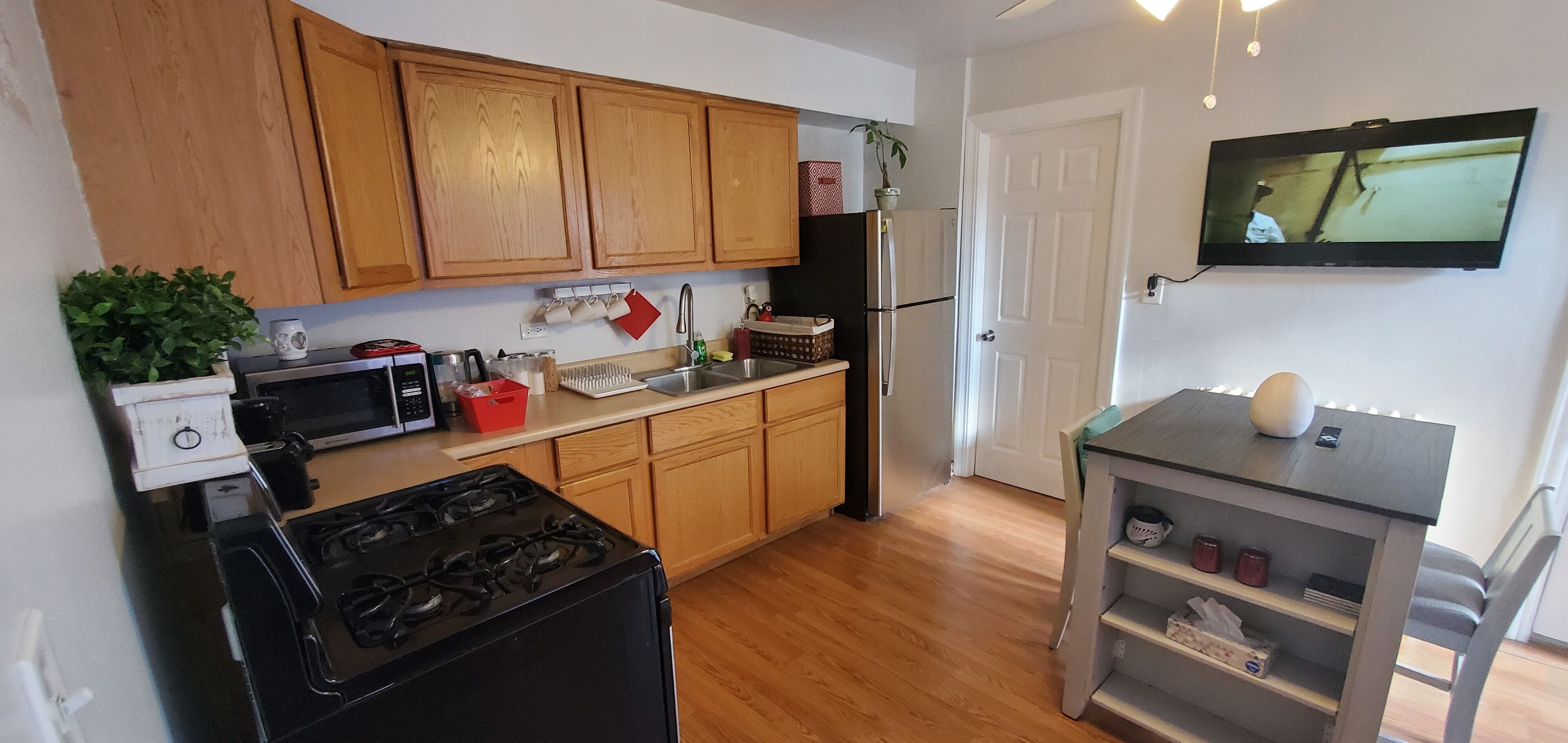 Una cocina compacta con armarios de madera, una estufa, un microondas, un refrigerador y una mesita con un florero.