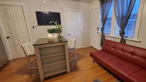 Een woonkamer met een rode bank, een eettafel met stoelen, een potplant en een flatscreen televisie aan de muur.