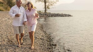 Una pareja caminando por una playa de guijarros con una masa de agua tranquila y árboles al fondo.