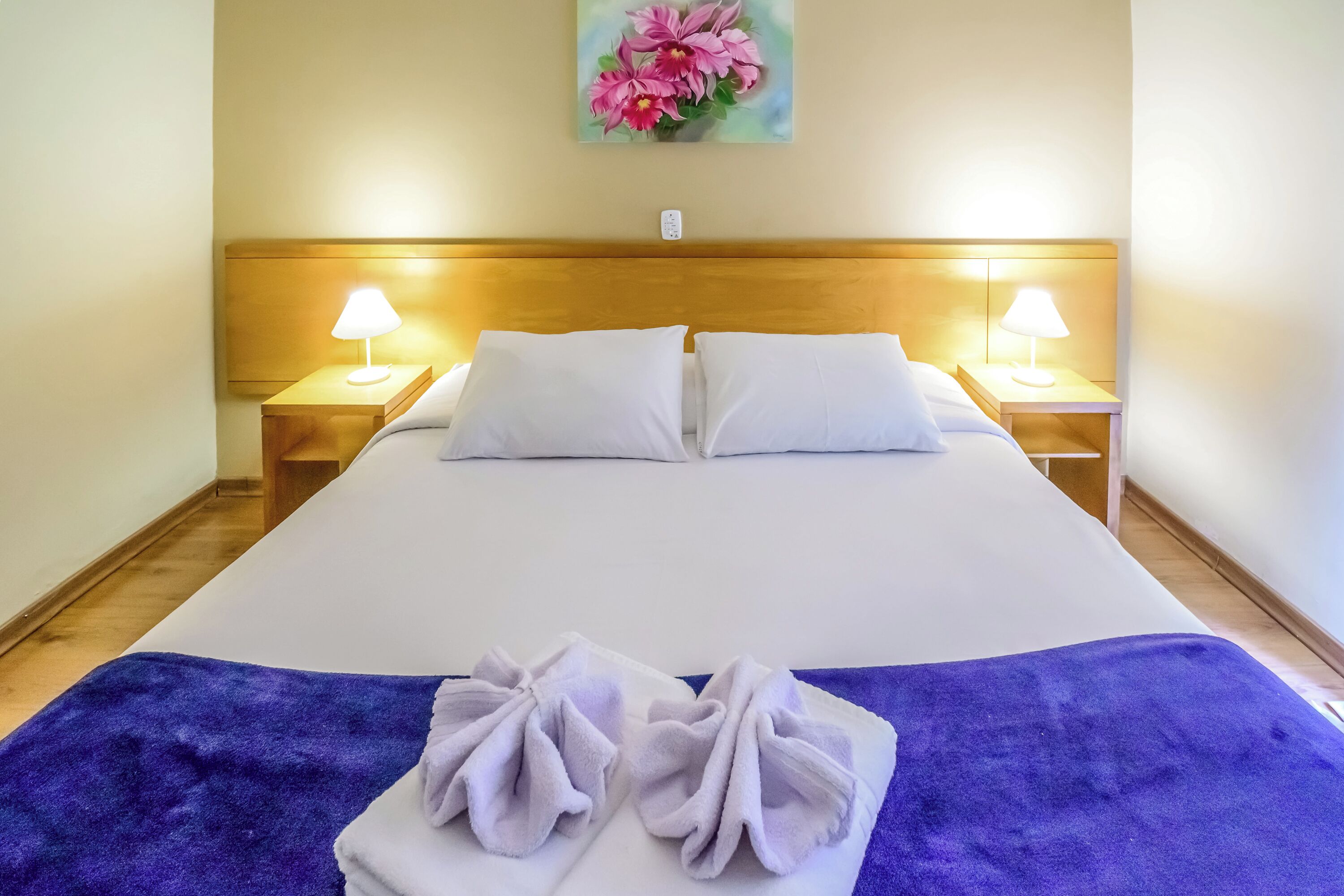 Une chambre d’hôtel avec un grand lit, deux lampes de chevet, une tête de lit en bois et un tableau représentant des fleurs accroché au mur.