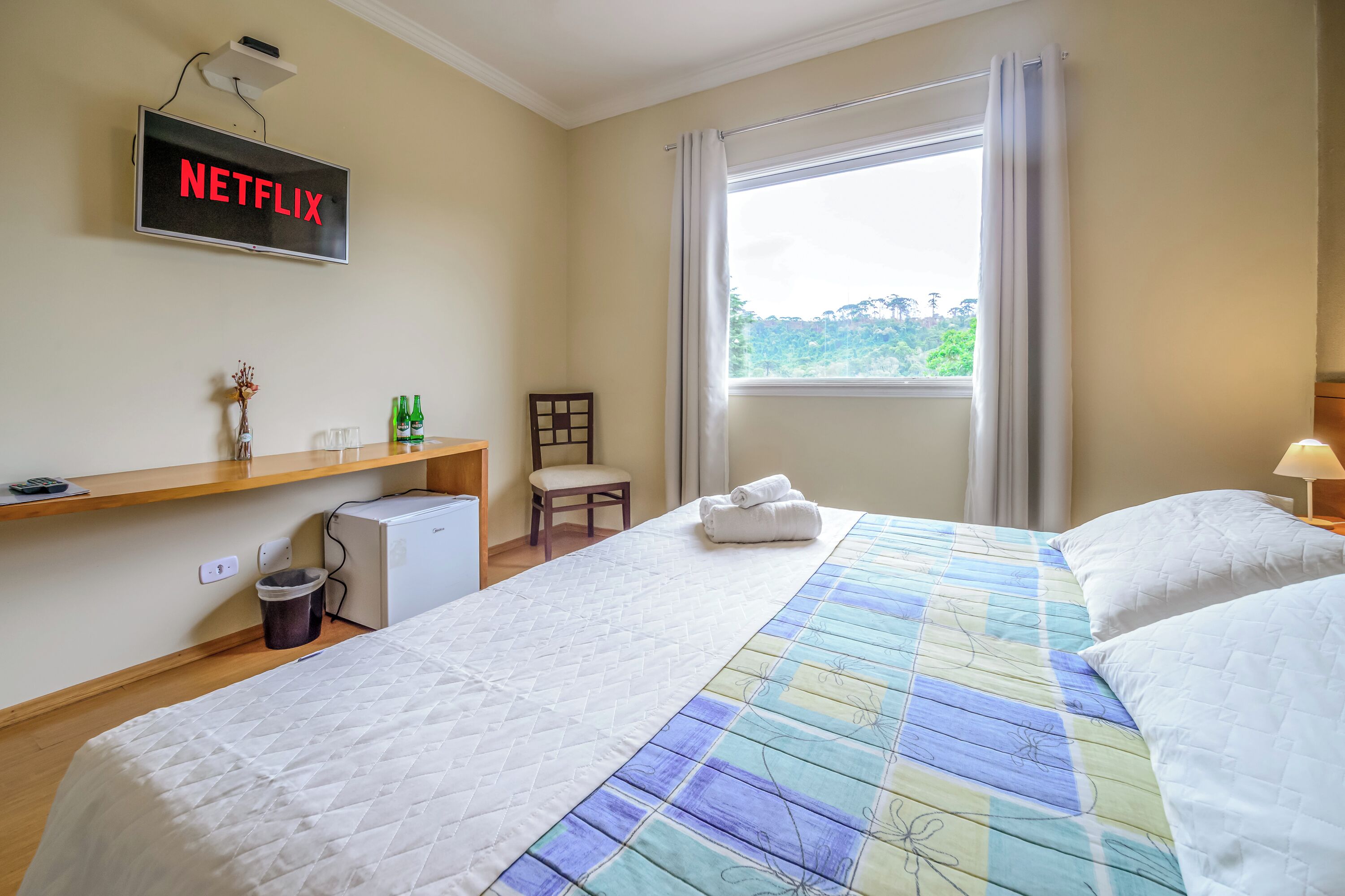A hotel room with a bed, a desk, a chair, a TV displaying 'NETFLIX', a window with curtains, and a view of greenery outside.