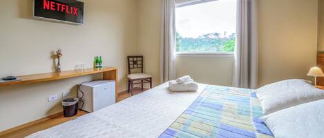 A hotel room with a bed, a desk, a chair, a TV displaying 'NETFLIX', a window with curtains, and a view of greenery outside.
