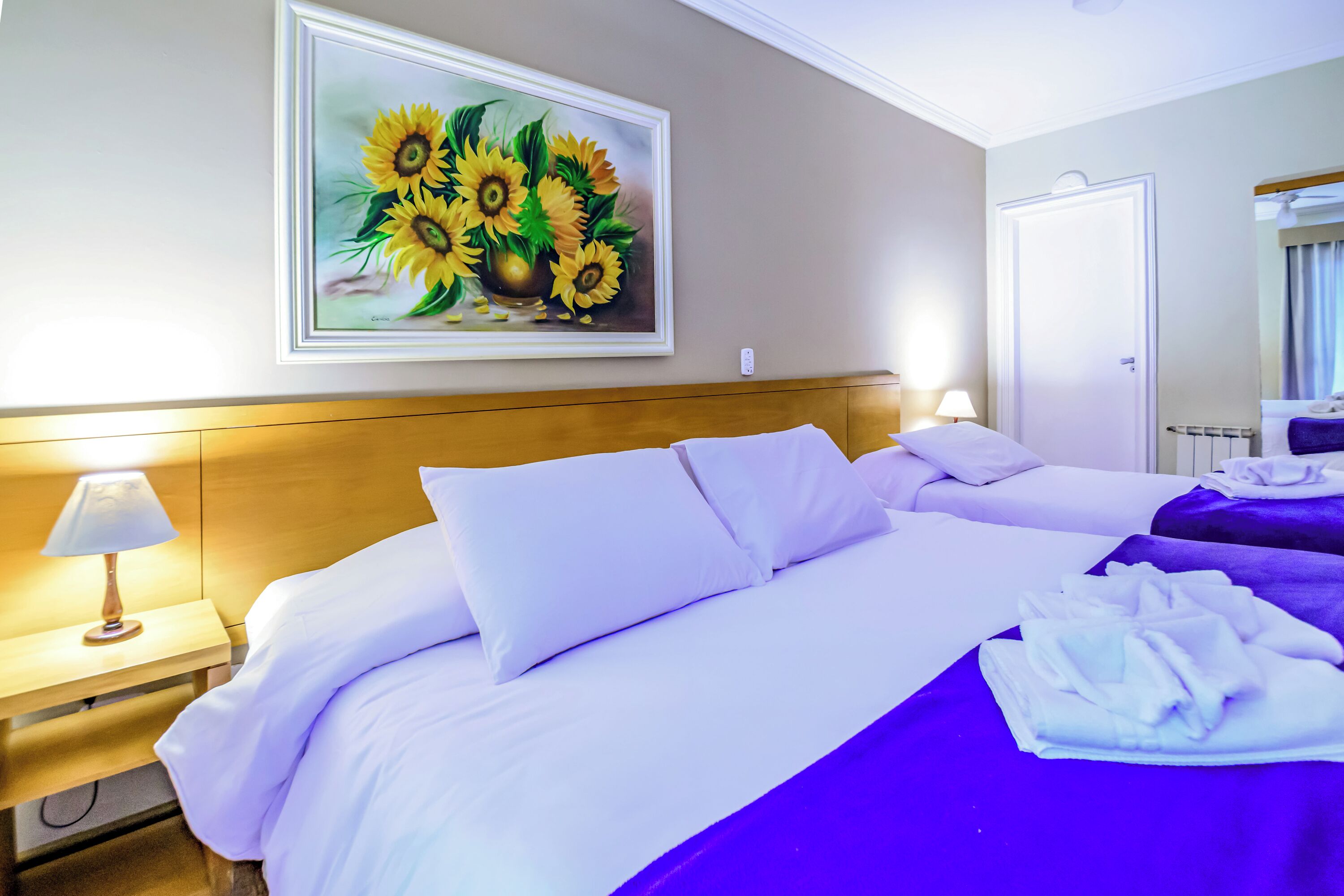 A hotel room with a large bed, a nightstand with a lamp, and a painting of sunflowers on the wall.