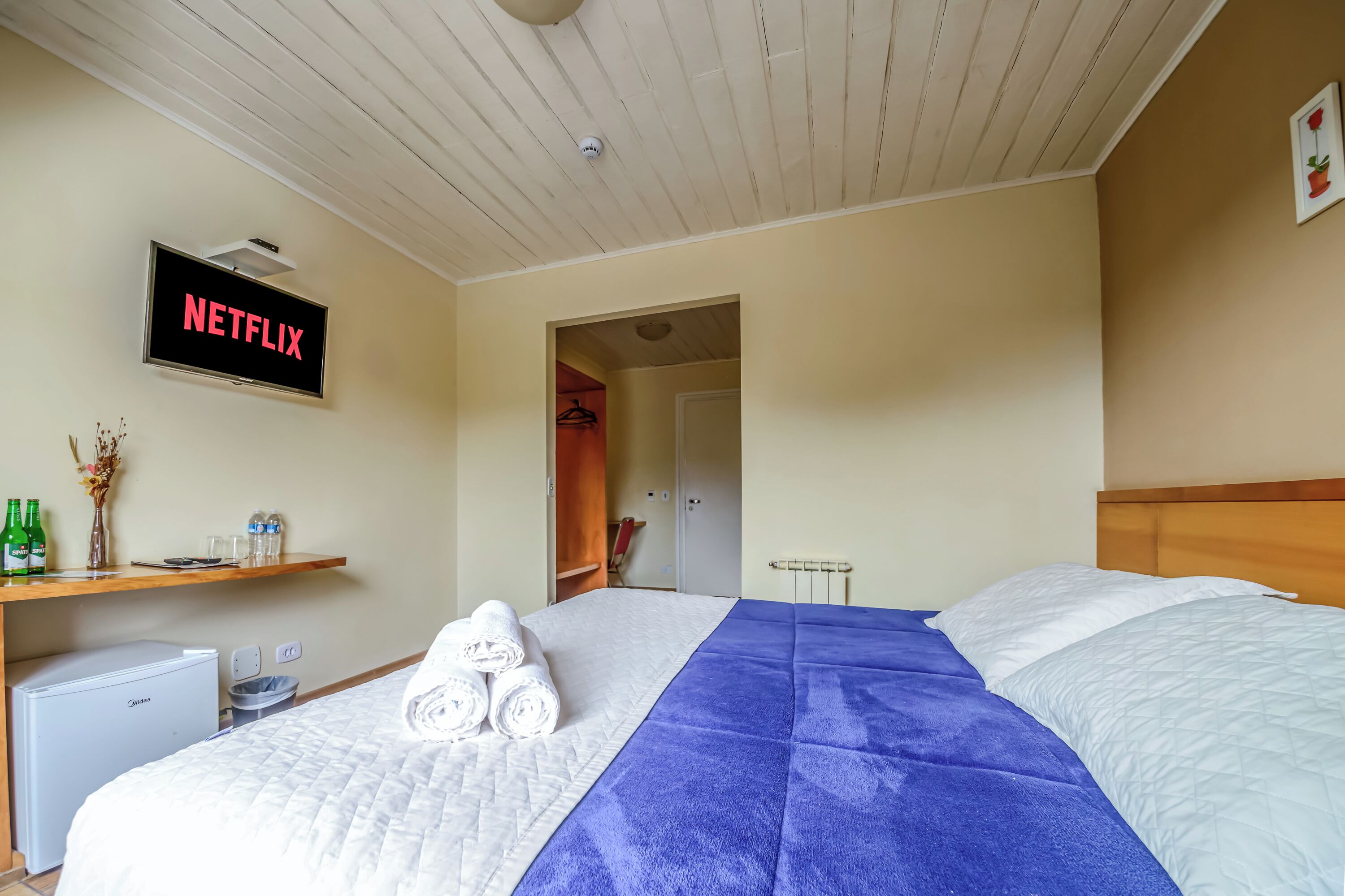 A hotel room with a bed, a small table, a TV displaying 'NETFLIX', and a refrigerator.