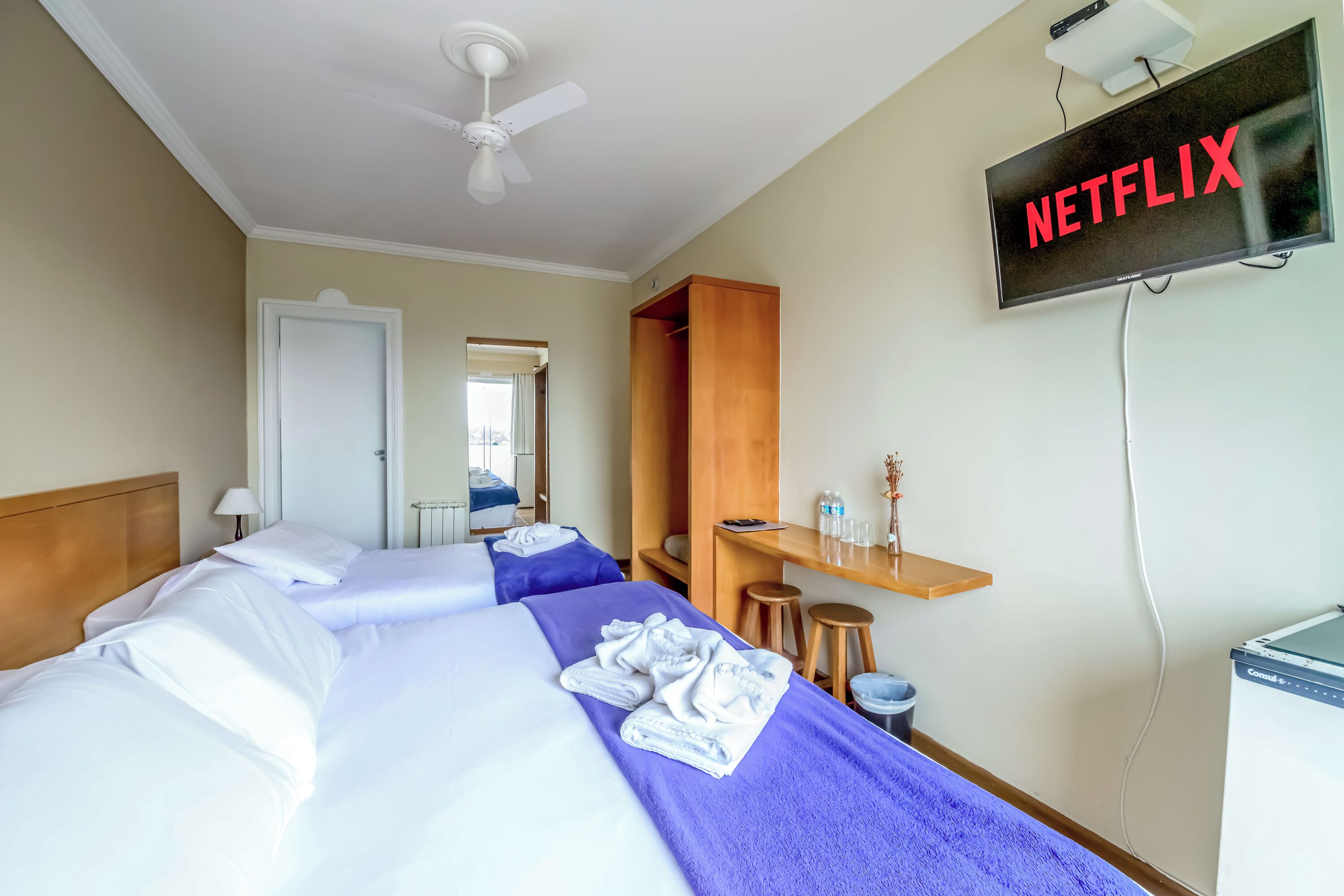 Une chambre d’hôtel avec deux lits, un ventilateur de plafond, un téléviseur affichant le logo « NETFLIX », une armoire en bois, une petite table avec une lampe et un mini-bar.