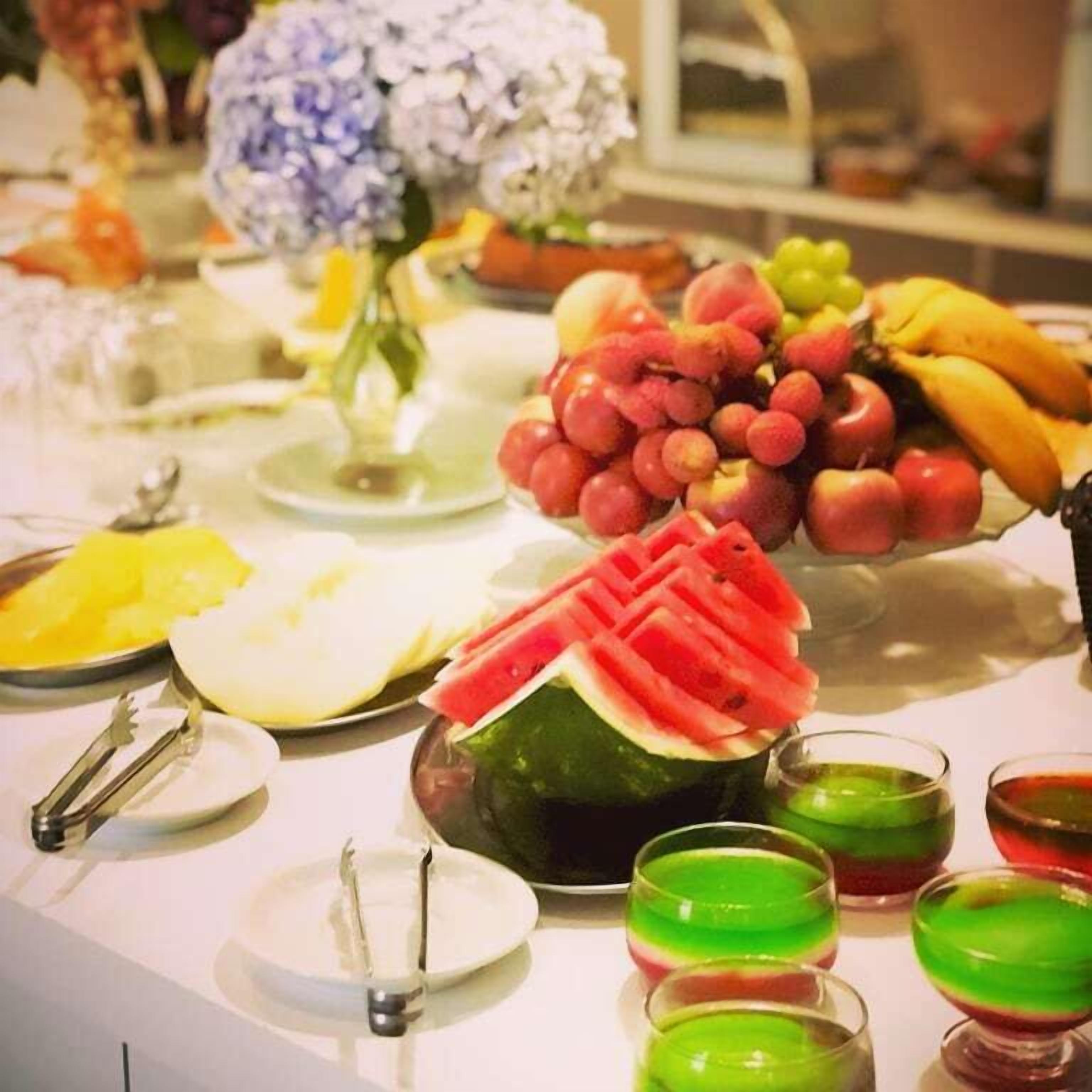 Een tafel met diverse soorten fruit, een watermeloen uitgehouwen in een decoratieve vorm, en verschillende glazen groene en rode drankjes.