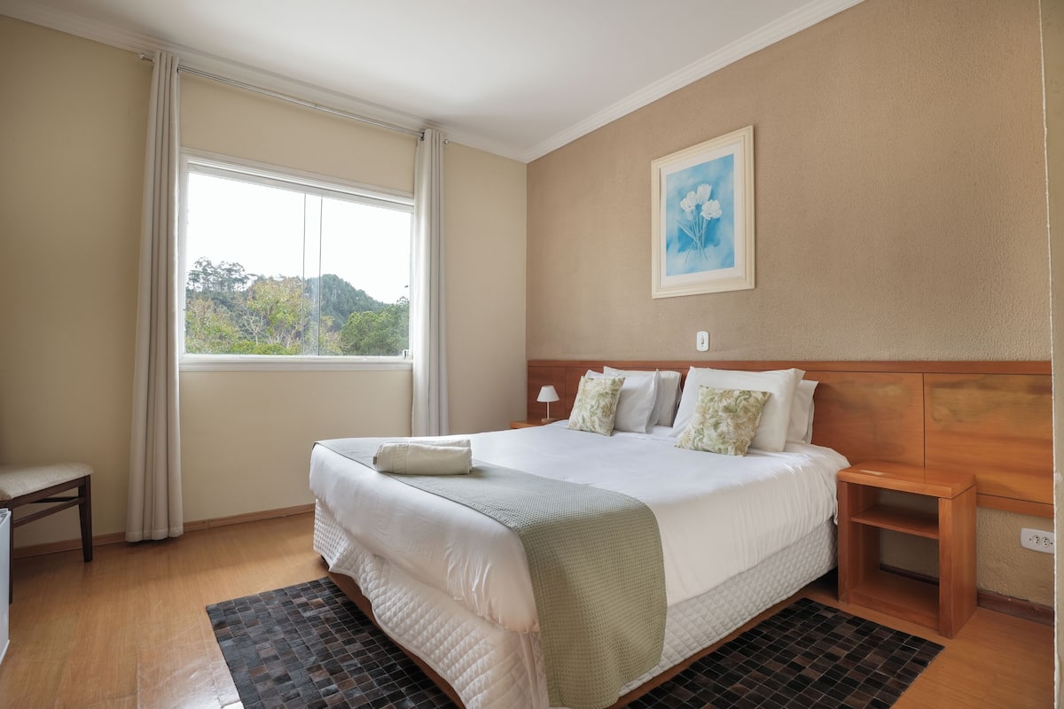 Habitación de hotel ordenada con una cama grande, una mesita de noche pequeña, una silla y una ventana con cortinas.