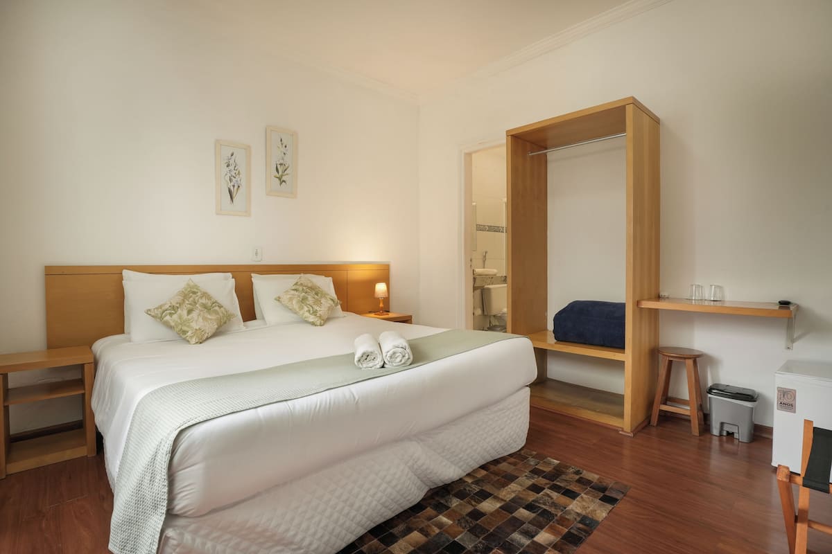 Habitación de hotel con una cama grande, cabecera de madera, dos mesitas de noche, un velador, un espejo, un taburete pequeño, un estante con vasos y un cubo de basura.