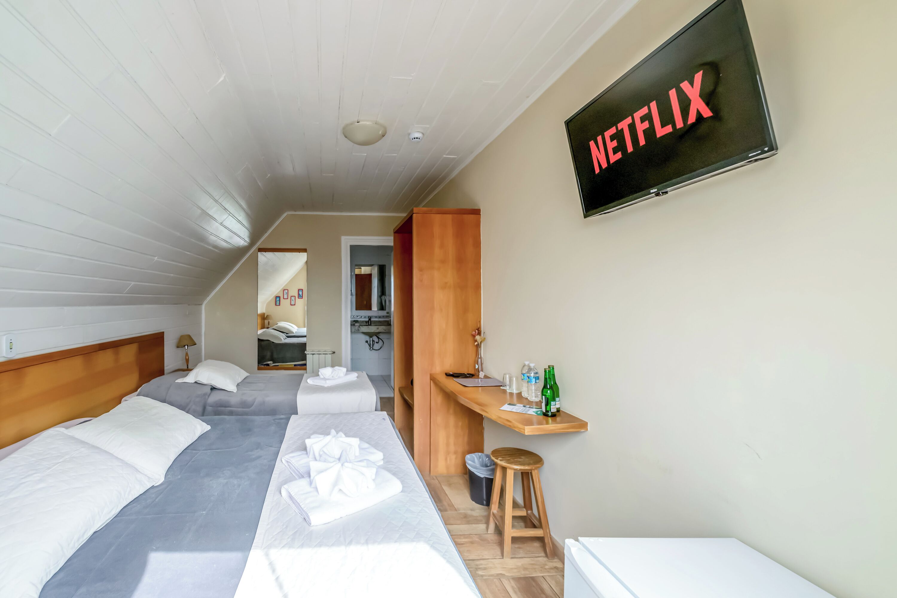 Habitación de hotel con dos camas, un escritorio de madera, un televisor montado en la pared y un taburete pequeño.