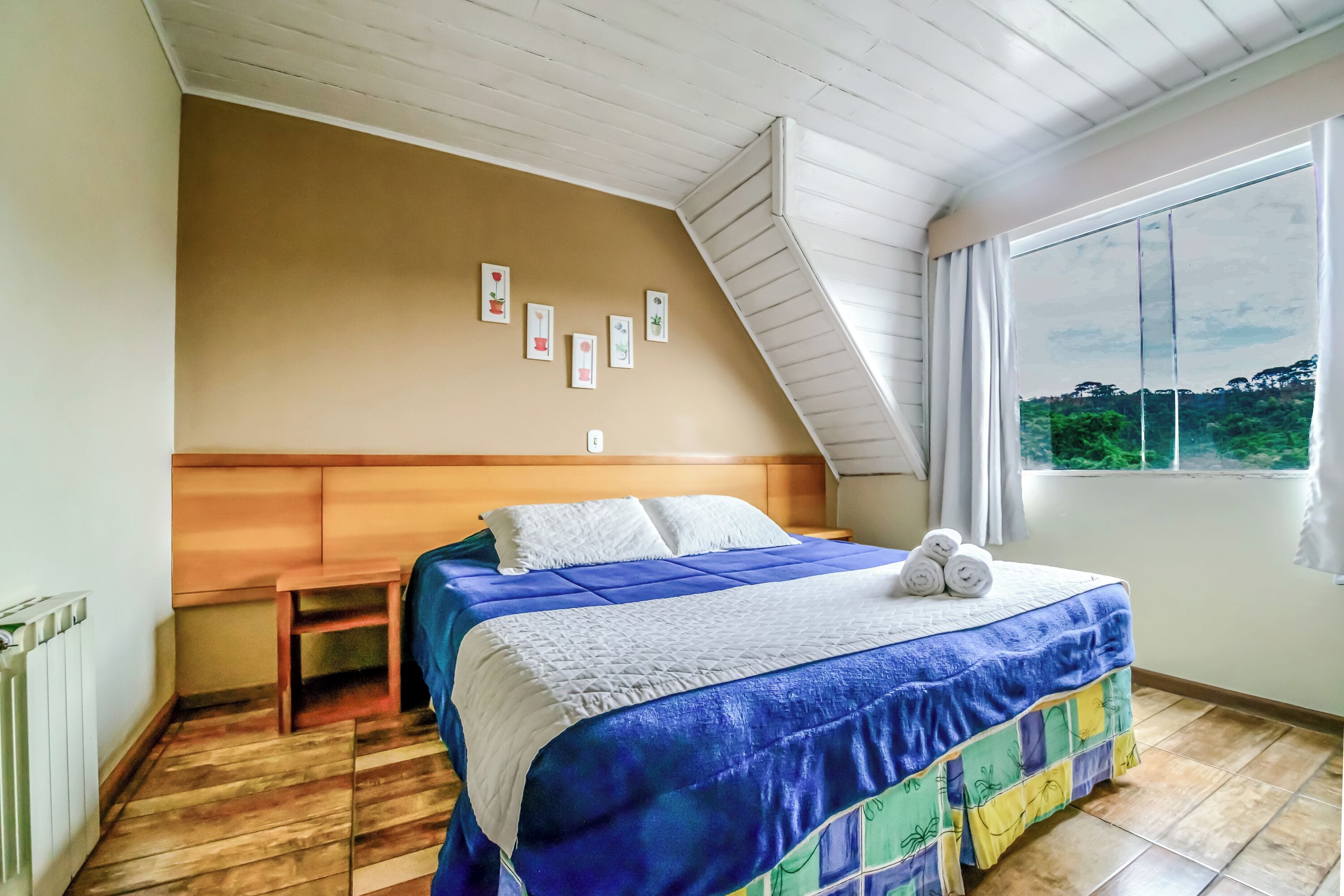 Un dormitorio con una cama, cabecera de madera, una banca, una ventana con cortinas y vistas a espacios verdes.