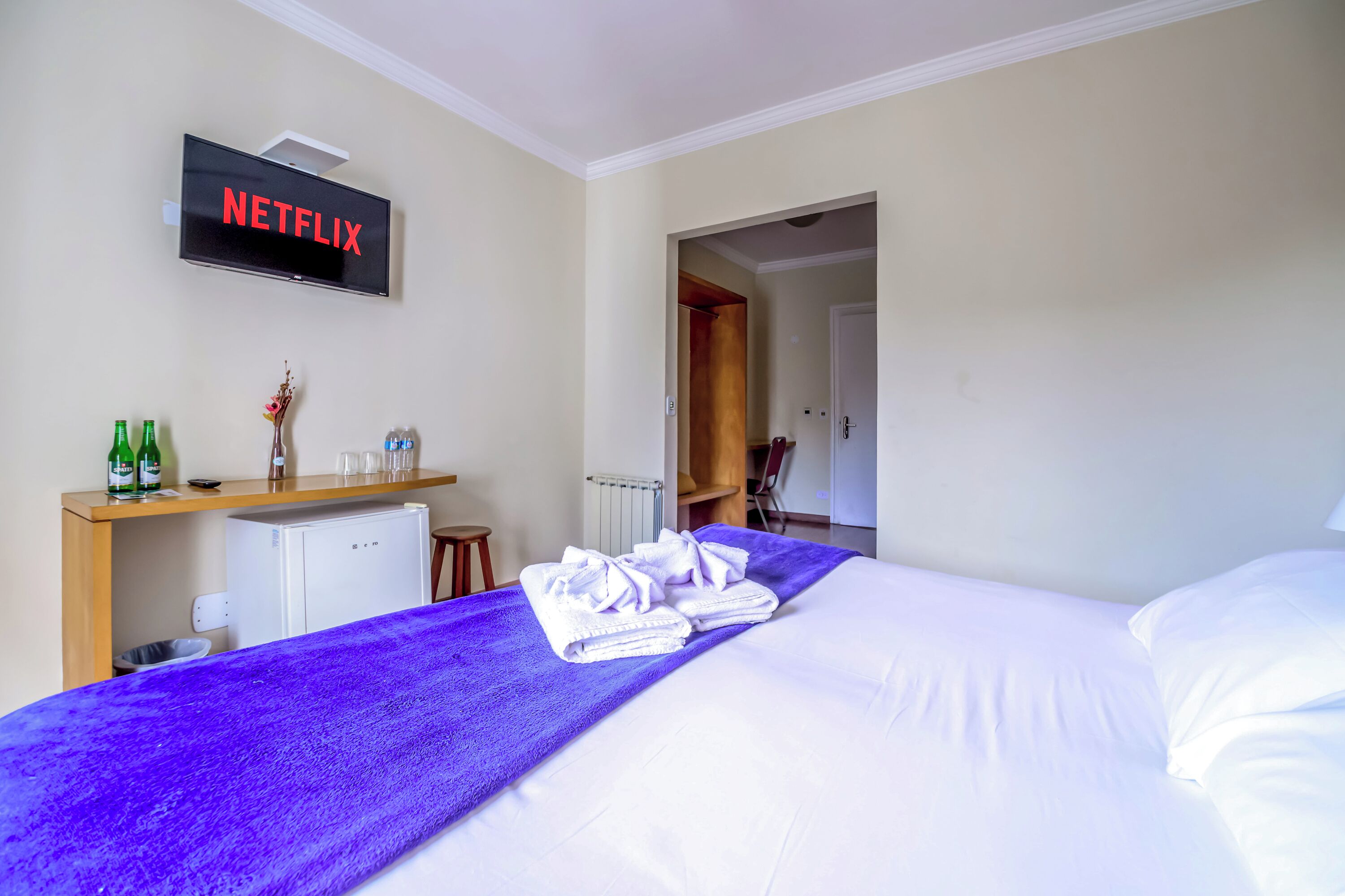 A hotel room with a bed, a TV displaying 'NETFLIX', a small fridge, a wooden shelf with bottles and glasses, and a bathroom door visible in the background.