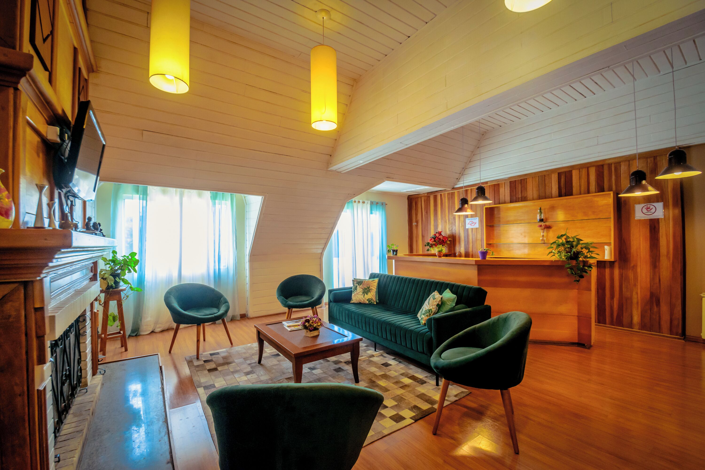 Una sala de estar acogedora con un sofá verde, dos sillones, una mesa de centro, suelo de madera, una chimenea y una zona de cocina con armarios de madera y luces colgantes.
