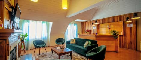 Una sala de estar acogedora con un sofá verde, dos sillones, una mesa de centro, suelo de madera, una chimenea y una zona de cocina con armarios de madera y luces colgantes.