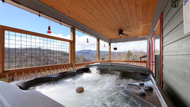 A hot tub with a wooden deck and large windows overlooking a mountainous landscape. - 3BR 2.5BA Mnt Views! Hot Tub/Pool Table! 2mins to downtown! (Gatlinburg)