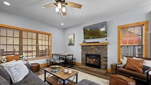 A spacious living room with a fireplace, large windows with blinds, a ceiling fan, and a television mounted above the fireplace. - 3BR 2.5BA Mnt Views! Hot Tub/Pool Table! 2mins to downtown! (Gatlinburg)
