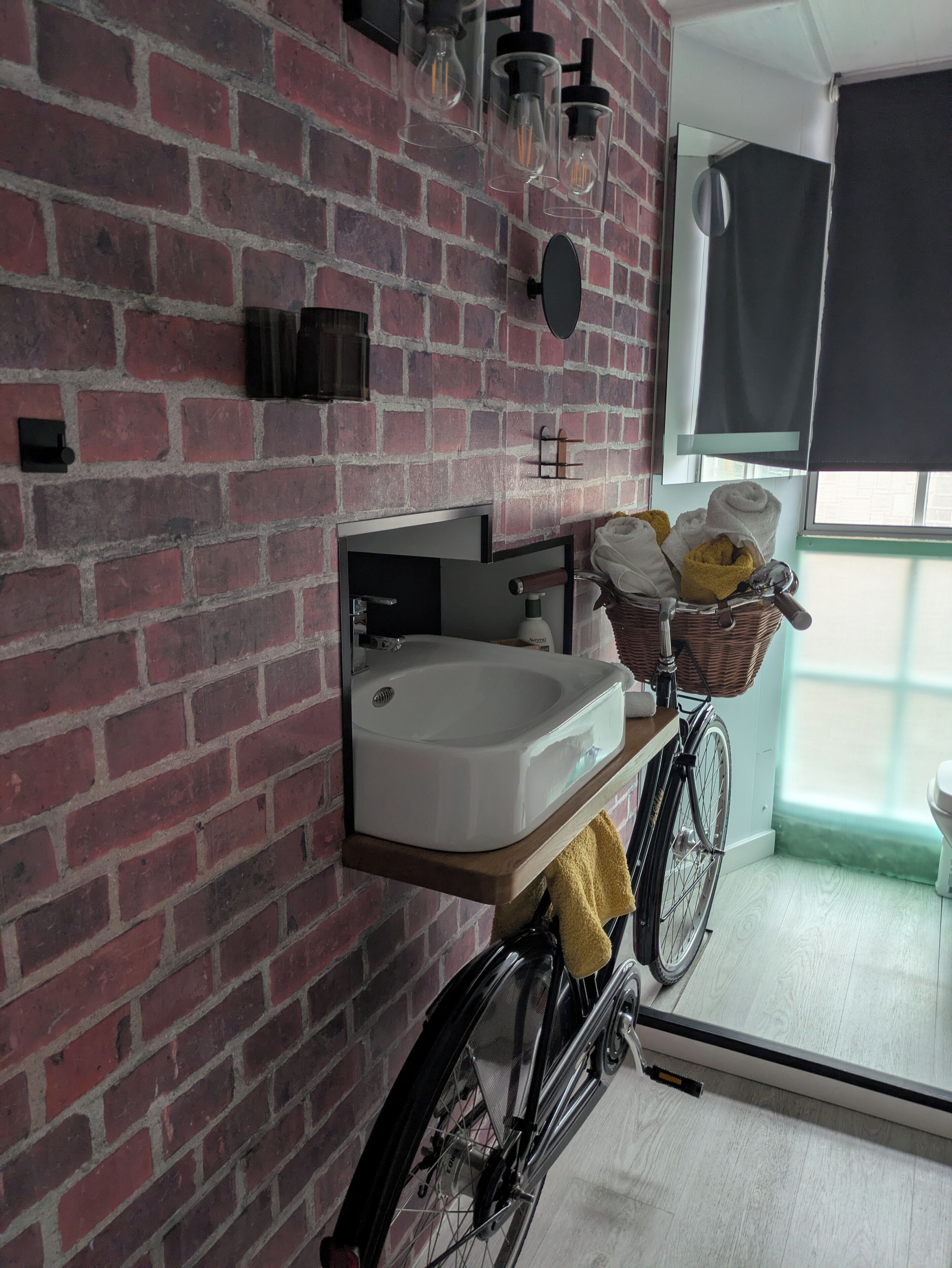 Et badeværelse med en murstensvæg, en hvid vask, et spejl og et cykelstativ med håndklæder.
