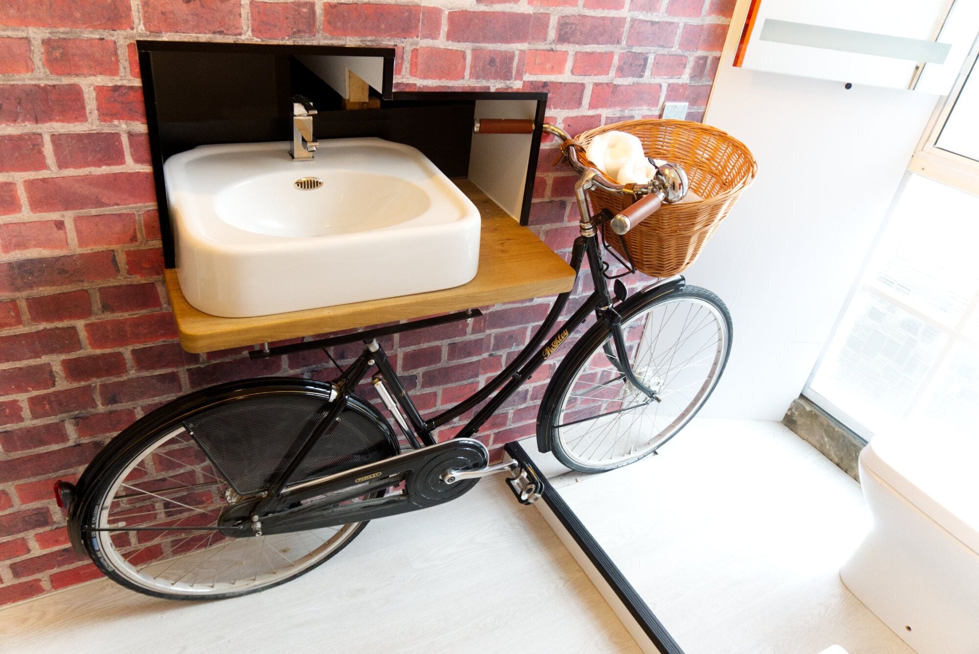 Un vélo avec un panier fixé à l’avant, garé contre un mur de briques, avec un évier blanc monté sur une étagère en bois.