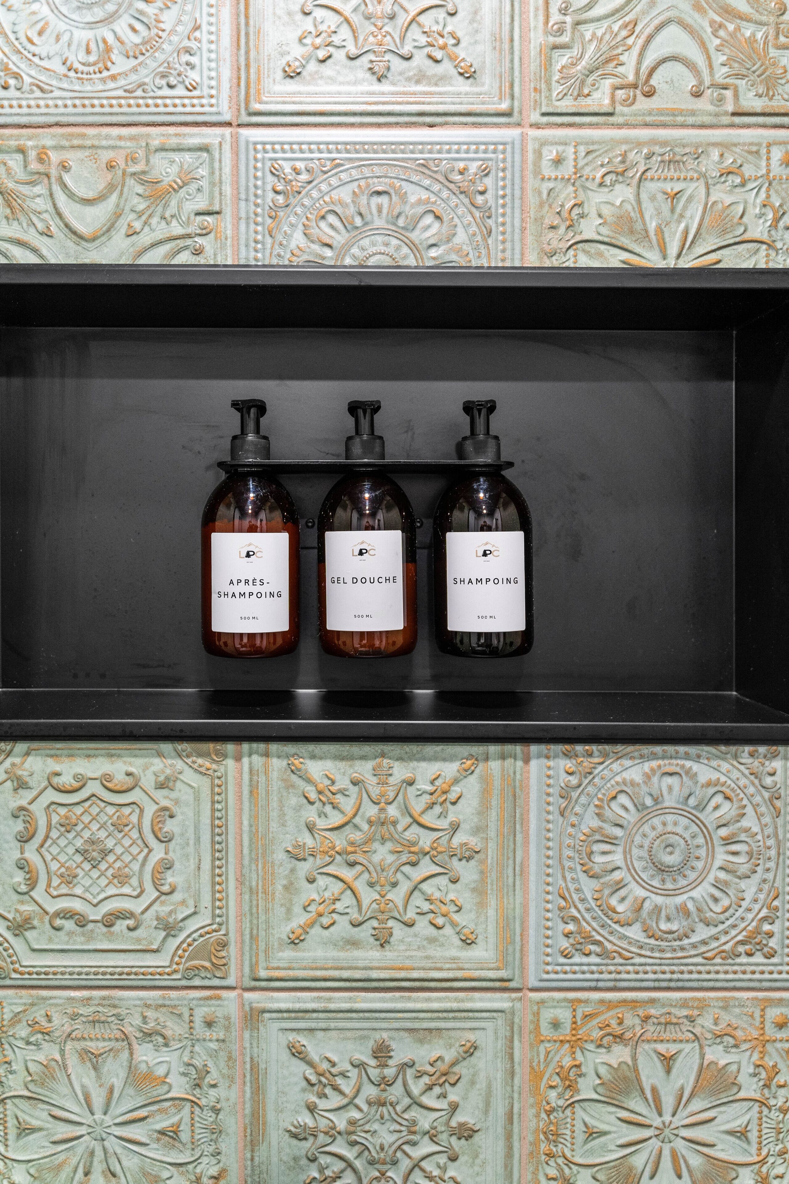 Une étagère avec trois bouteilles de shampoing, le tout placé devant un mur décoratif recouvert de carreaux.