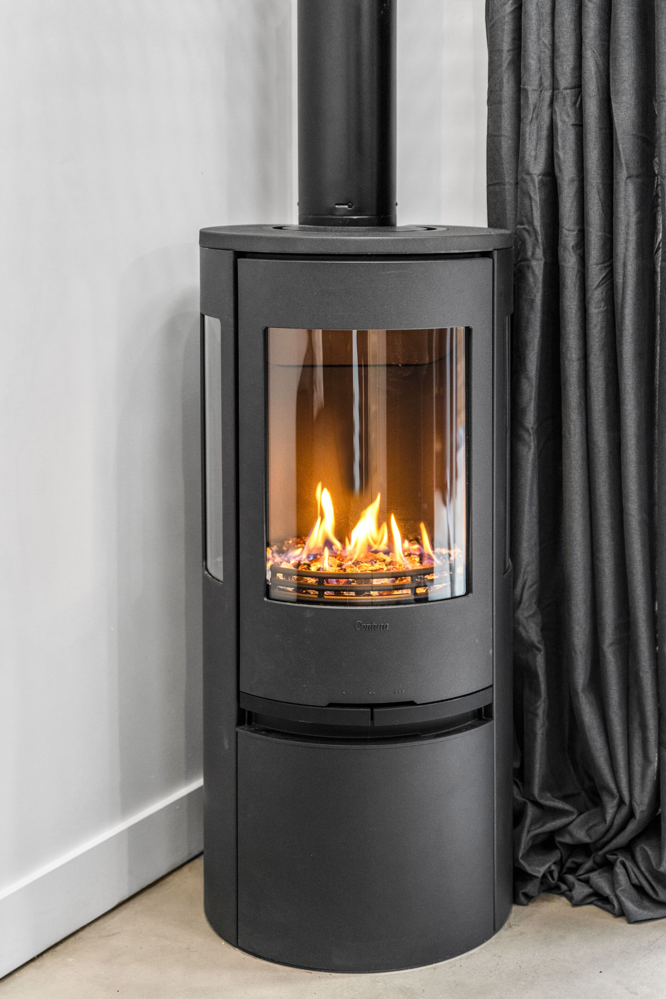 A modern, black wood-burning stove with a glass front, positioned against a white wall and black curtains.