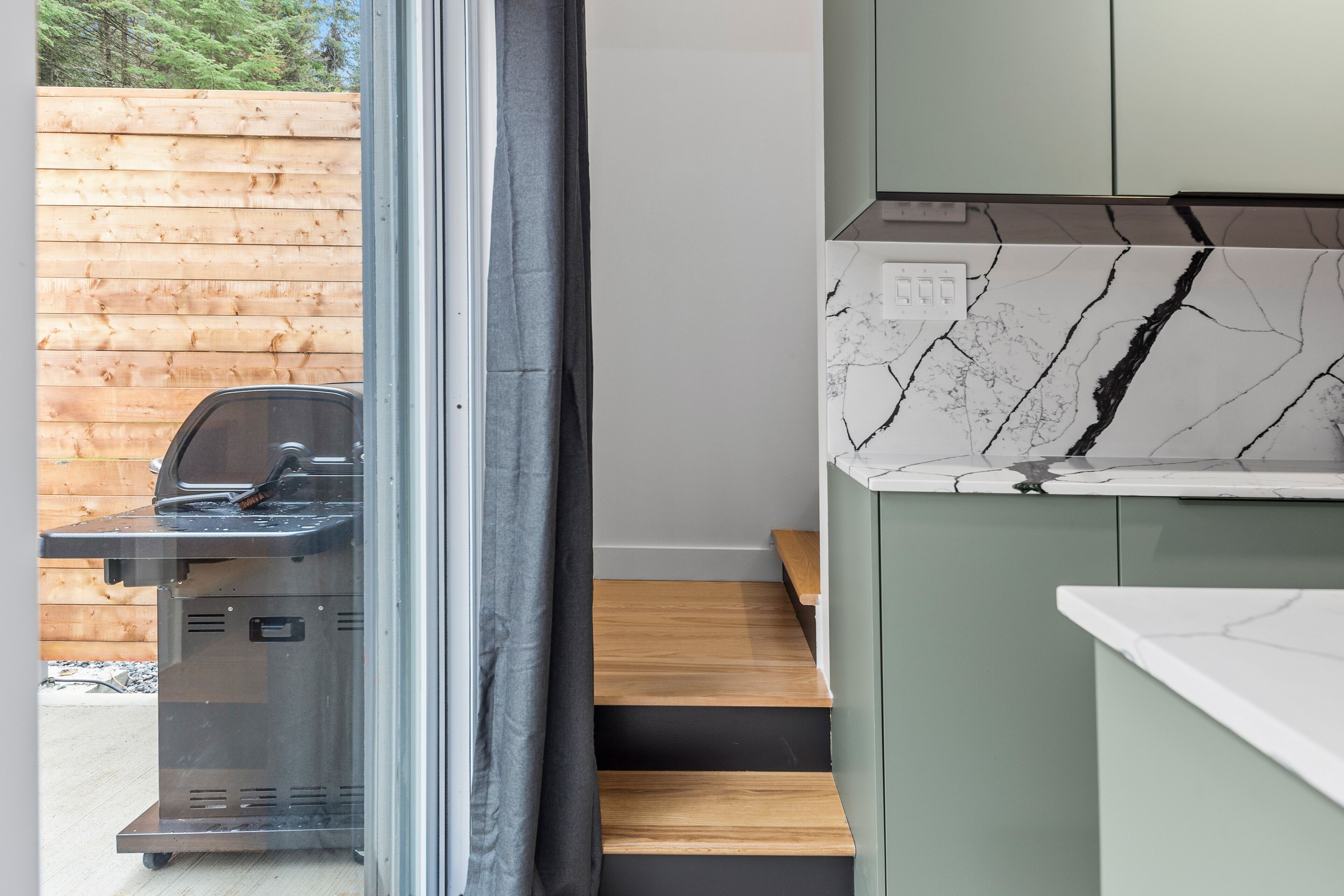 A modern kitchen with a marble countertop, wooden steps, and a barbecue grill outside.