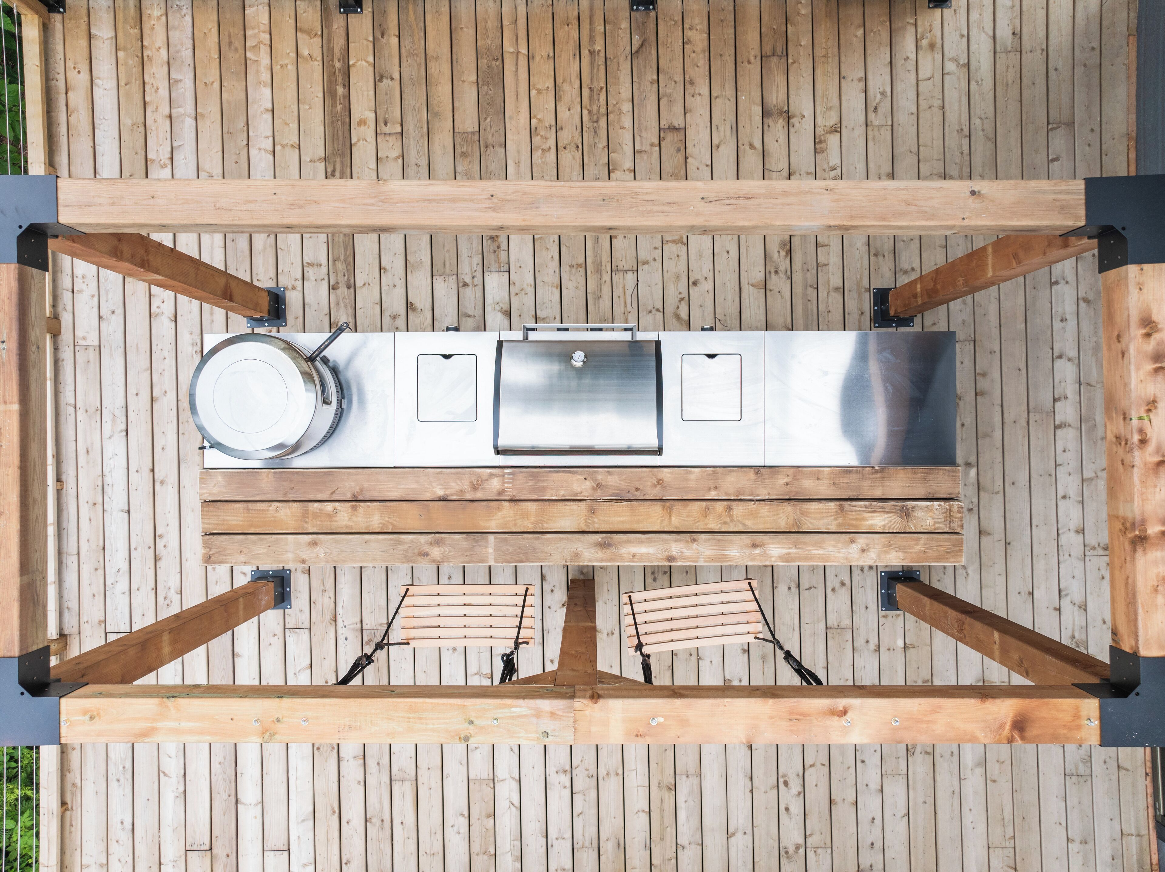 A top-down view of a wooden deck with a stainless steel grill station, two wooden chairs, and a metal pot.