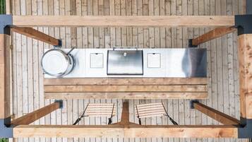 A top-down view of a wooden deck with a stainless steel grill station, two wooden chairs, and a metal pot.
