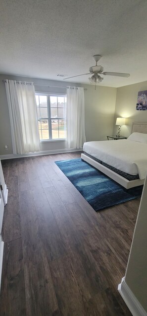 Quarto com cama, ventilador no teto, janela com cortinas e mesa de cabeceira com abajur, além de vista para o exterior.