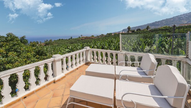 A terrace with white metal chairs and a white balustrade overlooking a lush garden and distant hills.