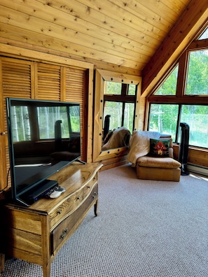 Ein gemütliches Zimmer mit Holzboden, einem großen Fenster, einem Fernseher auf einer Holzkommode und einer bequemen Sitzecke.