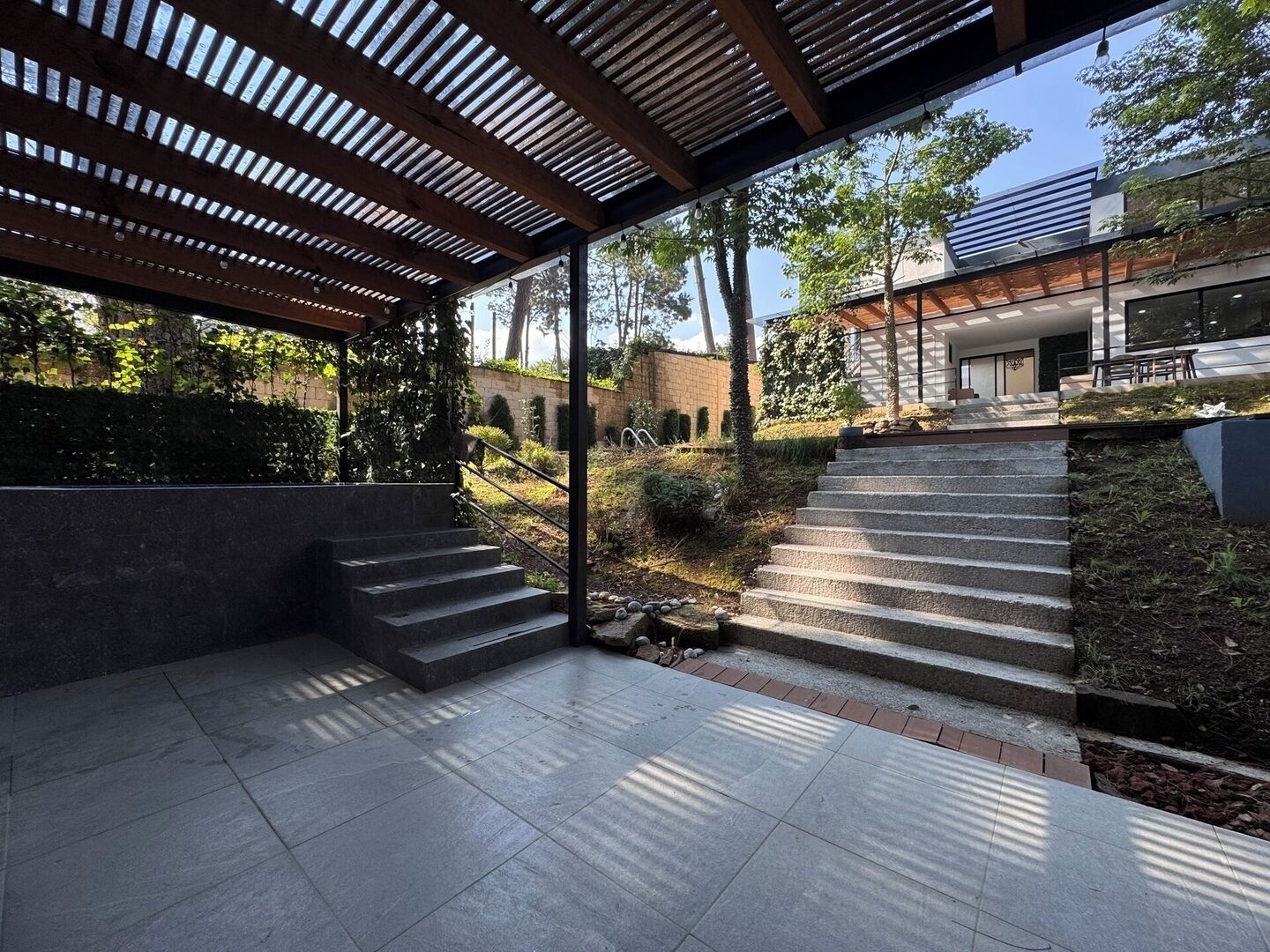 Un espacio moderno al aire libre con un porche cubierto, escalones de piedra y vista a un edificio.