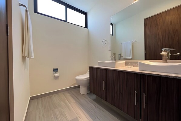 Une salle de bain moderne dotée d’un grand miroir, de deux lavabos, de toilettes et d’une fenêtre laissant entrer la lumière naturelle.