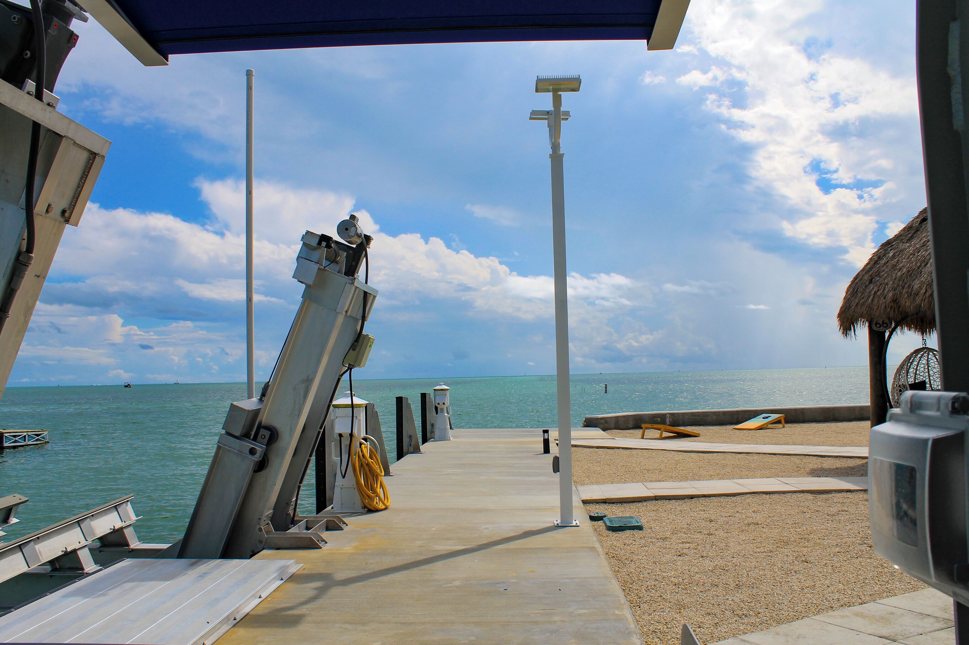 A pier with a concrete walkway leading to a beach, a thatched-roof structure, and a lifebuoy.