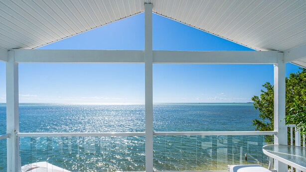 Un balcón techado con vistas despejadas al mar, barandilla de cristal y un banco blanco.