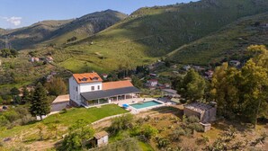 Ein modernes Haus mit Swimmingpool, umgeben von üppigen, grünen Hügeln und wolkenlosem Himmel.