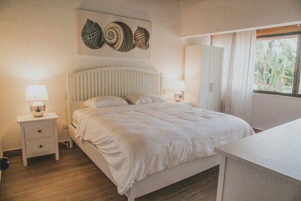 A bedroom with a bed, bedside tables, a lamp, a wardrobe, and a window with a view of greenery. - Cozy Entire Villa with Picuzzi (La Romana)
