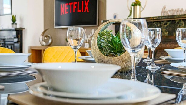 A dining table set with plates, bowls, and wine glasses, with a television displaying "NETFLIX" in the background. - Thrift House • Central Location • Pool Table • Free Parking • Smart TV • Fast WiFi • Yoko Property (Northampton)