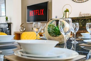 Une table à manger est dressée avec des assiettes, des bols et des verres à vin, et un téléviseur affiche le mot « NETFLIX » en arrière-plan.