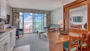 A compact hotel room with a kitchenette, a dining area, a sofa, and a balcony with a view of the sea.