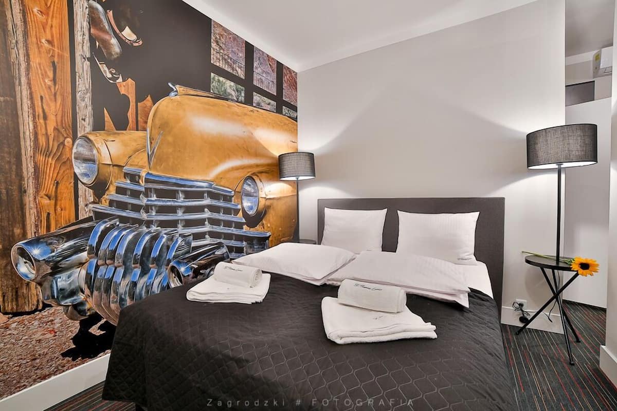 Habitación de hotel con un gran mural de un coche antiguo, una cama con toallas ordenadas, una lámpara de noche y una pequeña mesa con un girasol.