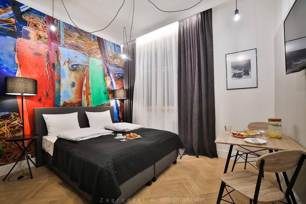 Una habitación de hotel moderna con una cama grande, una mesa de comedor con desayuno y un cuadro abstracto colorido en la pared.
